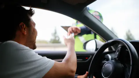 Stock image taken inside a car of a driver handing his licence to a police officer. The driver has short dark hair and is wearing a white T-shirt. The officer is blurred by distance and partly unseen due to the frame of the car.