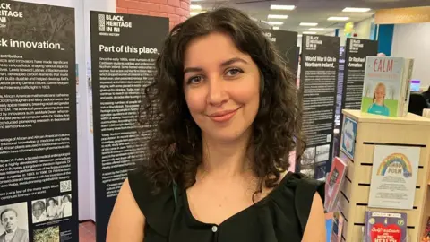 Aynaz Zarif has long brown hair and is wearing a black dress. 
She is standing in a library with books in the background and also standing close to the exhibit. 