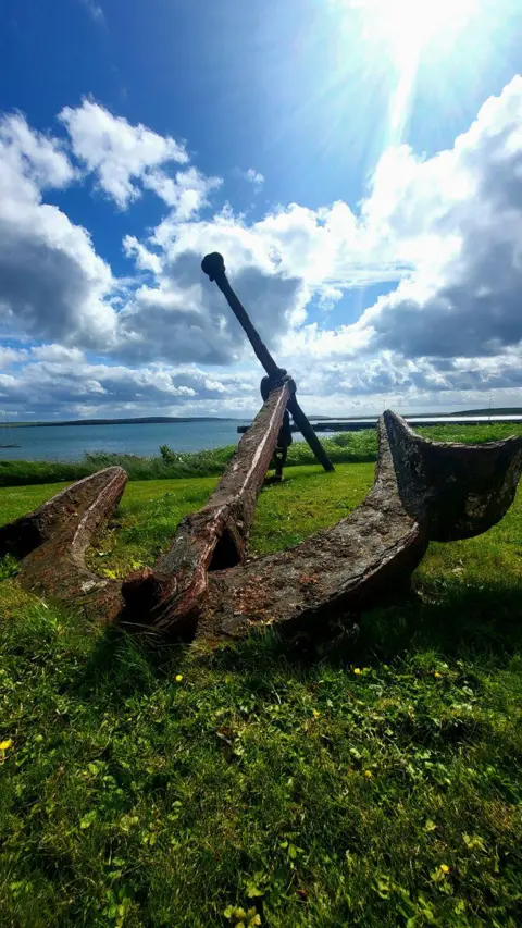 Jessica Bruce An old anchor against a skyline with sun breaking through the clouds