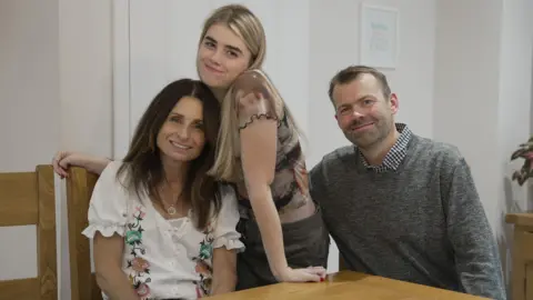 A teenage girl with long blonde hair stands between her mother and father who are sitting at the kitchen table. All three are smiling and facing the camera.