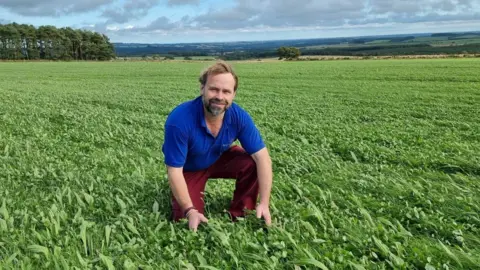 Simon Bainbridge Simon Bainbridge, wearing red trousers and a blue top, is kneeling in a field of pasture with hills behind. 