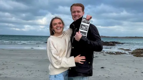 Image shows athlete Alastair Chalmers and his partner on a beach, holding a photo scan of their unborn baby. Chalmers, who has short ginger hair, wears a dark-coloured coat, his partner is pictured in a cream jumper, with jeans and long blond hair. They are both smiling and pictured amidst a backdrop of a choppy sea, rocks and dark grey clouds overhead. 