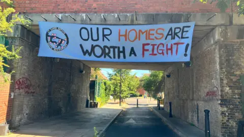 A large banner hangs on a railway bridge that crosses a pathway in suburban London. 
