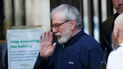 Adams is wearing a navy fleece top and is holding is hand up towards the camera. He has grey hair and a grey beard.