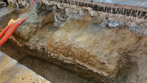 Museum of London Archaeology A side view showing several layers of the modern tarmac surface of Old Kent Road, with layers of roman road underneath. Orange tubes are visible to the left of the cross-section