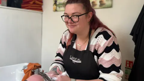Hairdresser Courtney, wearing an apron standing over a sink washing another person's hair in a salon. Shelves, folded towels, and wall art are visible in the background.