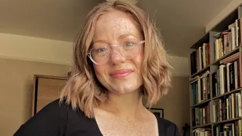 Laura Etchells A smiling Laura Etchells wearing glasses and smiling, standing in her living room with bookshelves seen on the right