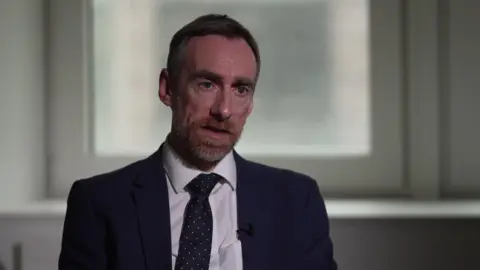 Detective Chief Superintendent Craig Nicholson wears a dark suit and tie while sitting in front of a window.