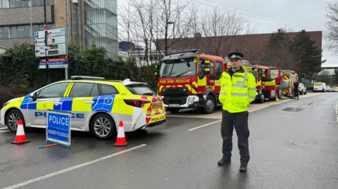 A police officer stands with a hi-viz jacket on. There is a police car and a police road closed sign and some traffic cones. Two fire engines are also parked next to each other.