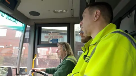 Three refuse workers, Dave, Greg and Dan, sit in the lorry with Dave driving and the other two looking out the windows.