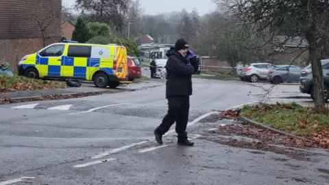 Dawid Wojtowicz/ BBC A police officer in black coat, hat and trousers walks across a road on the phone. A police van and police tape is across the road.