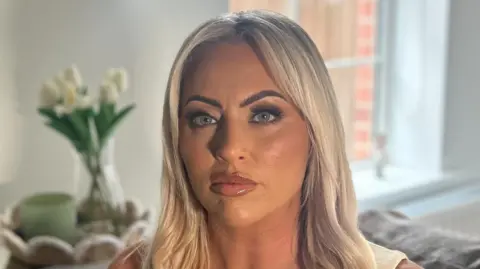 A woman with long blonde hair and blue eyes is looking directly into the camera, unsmiling. She is wearing a light-coloured sleeveless v-neck top. The background is out of focus but she is sitting on a grey sofa with light green cushions, behind her is a table with white flowers on it and a window. 