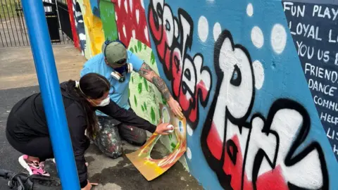 A male and female artist spray paint a wall. The words Spider Park are among the work on the wall.