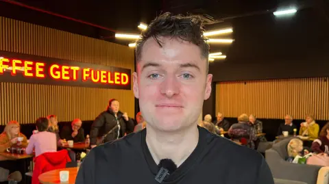 Joe wears a black t-shirt and is smiling, he's sat with his coffee shop behind him, where tables have customers sat at them. In the left of the show, a sign reads "Coffee Get Fueled".
