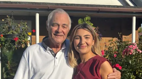 Hanan and her grandad embrace side by side for a picture in a garden on a sunny day. He has short white hair and wears a white T-shirt. She has long blonde hair and wears a red sleeveless top. Hanging baskets with flowers are in the backdrop.