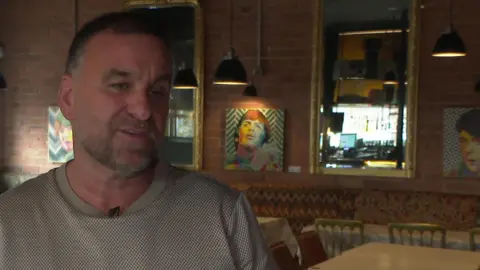 John Currie with short grey hair and beard wearing a beige and white checked t-shirt. He is standing in an empty restaurant.