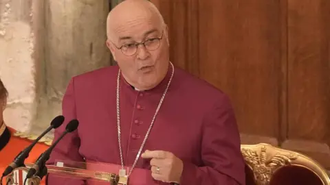 The Most Reverend Stephen Cottrell, who is wearing a burgundy gown with a gold cross around his neck, speaks during a dinner service.
