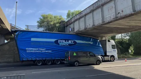 A white cab and a blue trailer on a road. The articulated lorry's trailer is stuck under a grey bridge.