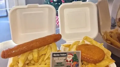 Natassa Trattou There is a takeaway tray witha jumbo sausage and chips on the left and a battered fish cake and chips in a takeaway tray on the right.