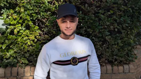 Clayton wearing a white long-sleeved top that has the word 'Glorious' written in gold letters. He stands in front of a bush.