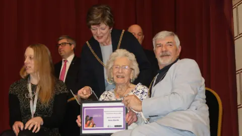 West Northamptonshire Council Six people are in the picture, and appear to be on a stage. An older woman is sat down in the centre, holding some sort of certificate. A woman with a mayoral chain is leaning over her, standing behind. A red velvet curtain is in the background.