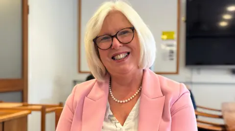 Dr Nicola Brink is wearing a pink jacket as she sits in a room and smiling directly at the camera. She has pale blonde hair and glasses.