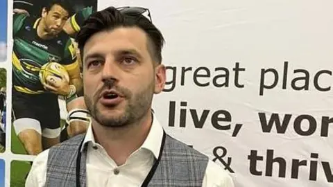 Oliver Conopo/BBC Thomas Manning with short dark hair and beard wearing a grey waistcoat and light-coloured shirt smiling at the camera.