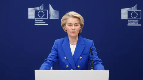 Reuters Ursula von der Leyen standing a podium speaking into a microphone. Behind her a blue background with the logo of the European Commission.