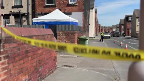 A residential street, with police investigation tape in the foreground. There is a police officer and cars in the distance.