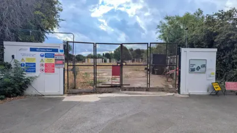 A field of concrete and dried, brown grass has been secured behing large metal gates and tempoary walls which have been painted white. The wall to the left of the gate is covered with construction site warning signs, some red, some yellow and some blue.