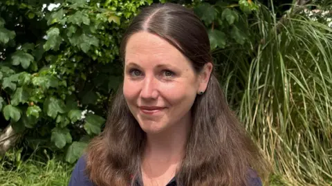 A young woman with long dark hair over both her shoulders wearing a dark blue top with flowers on it, standing in front of a hedge in a garden.