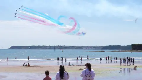 Crowds lining the beach as five planes fly next to each other in the sky with red white and blue smoke coming out behind.
