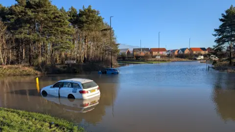 Sandy Lane Roundabout during the floods. Water covers the full three lanes of the roundabout. Two cars are partially submerged and stuck in the road. The water has covered part of the central roundabout and the roads leading from it. A housing estate can be seen in the distance.