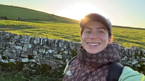 Chloe Cartwright takes a selfie while out walking in a thick brown North Face coat and a cap. She's smiling at the camera with a cobbled wall behind and a large open green field.