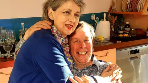 Handout A woman with short grey hair and a blue jumper embraces a man, wearing a black and white patterned shirt, as he sits on a kitchen chair. 