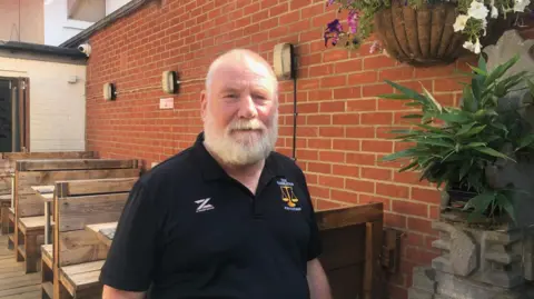 ALEX STEVANOVIC/BBC Andrew is standing in his pub garden.
There are seats behind him and a wall with plants next to him too.