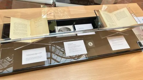Two books with accompanying information and pictures are in a large glass box.
They are stationed on a wooden table.