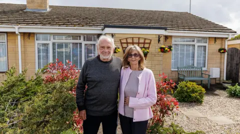 Anthony and Pamela stand in front of a bungalow made out of light stone. There is a neat garden, with gravel, small bushes and colourful hanging baskets. They both smile at the camera. Anthony has a beard and wears a dark grey jumper and dark trousers. Pamela has sunglasses on, a light pink cardigan and beige top.