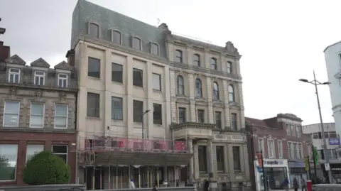 A building of at least four storeys is in the centre of the photo. Smaller buildings are next to it, either side, on the image.