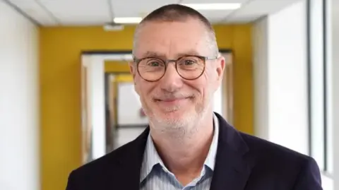 NUH A man with short grey hair and a beard and glasses. He is wearing a striped shirt and dark jacket