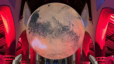 Cathedral Isle of Man A giant planet of Mars is suspended in a cathedral ceiling lit by red uplighting with large arches either side.