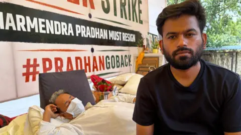 Geeta Pandey/BBC Abhijeet Dipke - wearing a black tshirt - looks into the camera, while Sonam Wagchuk lays down on a mattress in the background.