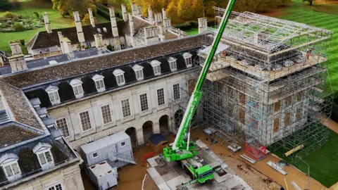 Conservation workers carrying out repairs on the roof of Boughton House, with extensive scaffolding in place. It features a large green crane, workers on the ground, The building is a very large stone building, with extensive windows. 