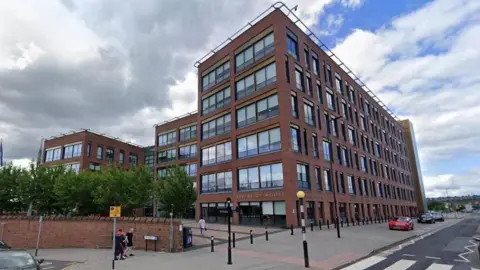 A six-storey building made from red brick. Several cars are parked to the right of it and a zebra crossing can be seen in the right corner of the picture.