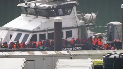 PA Media A group of people in orange hi-vis jackets disembarking a ship onto a port.
