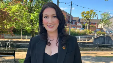 Little-Pengelly is smiling at the camera. She has shoulder length black hair and is wearing a black blazer and black dress. She is wearing a pin badge with a red poppy on it.