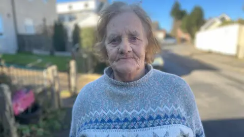 Doreen Tovey. She has short, fair hair and is wearing a grey, blue and white patterned jumper. She is pictured outside on Cobhorn Drive in Bristol. Houses and the road can be seen in the distance behind her. She is looking directly at the camera with a slight smile on her face