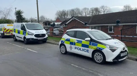 BBC Three police vehicles are parked outside a gated bungalow, with police cordon tape visible in the background