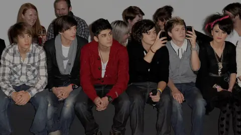 Getty Images Five young men and a young woman sit in the front row of a catwalk show. The group looks on with interest, with two of the male members holding up their phones to take pictures. The men are dressed in 2010-era fashion - a mixture of checked shirts, chunky cardigans and assymetrical haircuts. The young women uses a red Paisley bandana to tie up her curly black hair, and wears a selection of large gold pendants alongside her all-black dress and top combo. 