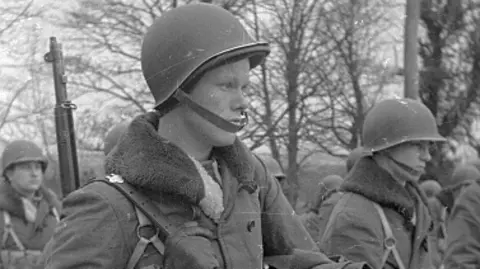 Haywood Magee Shows a black and white photo of US marines marching with hard helmets and trees in the background.